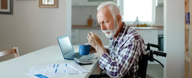 An elderly man in a wheelchair drinking coffee while working on a laptop at home.