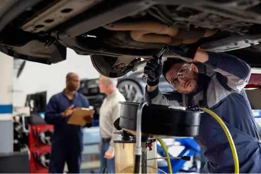 A man with protective eyewear stands under a raised car to perform maintenance on it while another man talks with a customer in the background