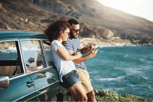 Man and woman leaning on a blue car on a nice day next to a body of water with hills in the background