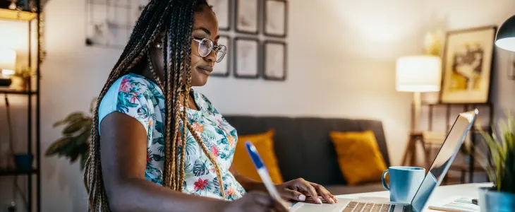 A woman in glasses works on a laptop and takes notes in a cozy home office.
