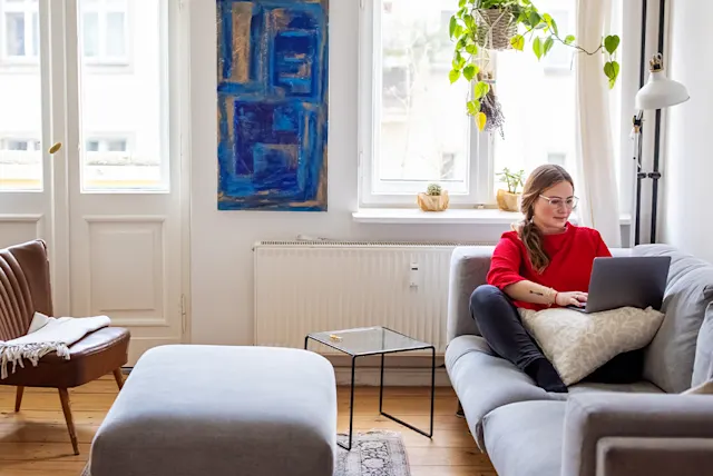 A women in a red sweater relaxes on a gray sofa, working on a laptop in a bright, modern living room.