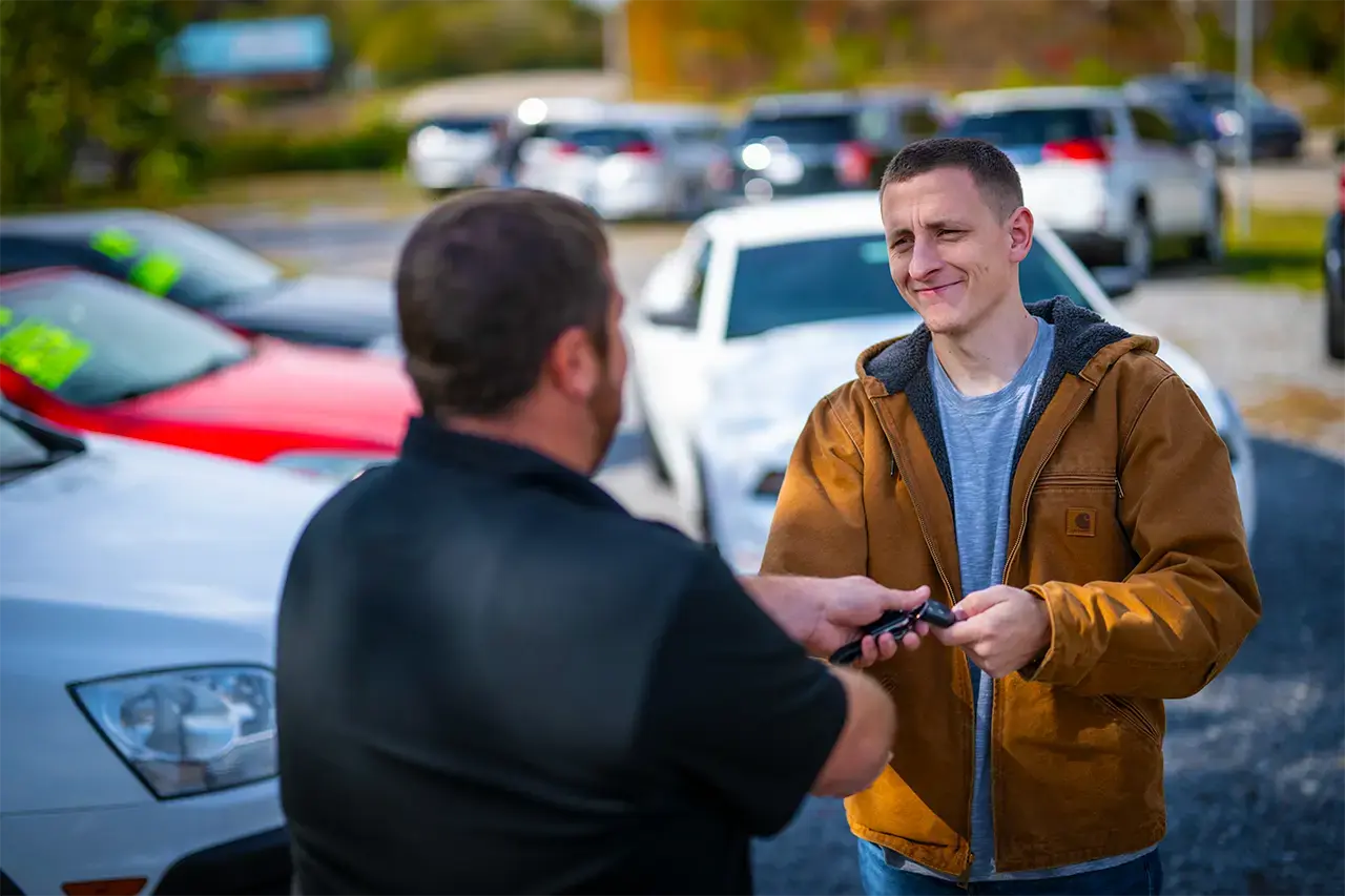 Two men in dealership parking lot with cars in the backing. The men are shaking hands and while exchanging card keys.