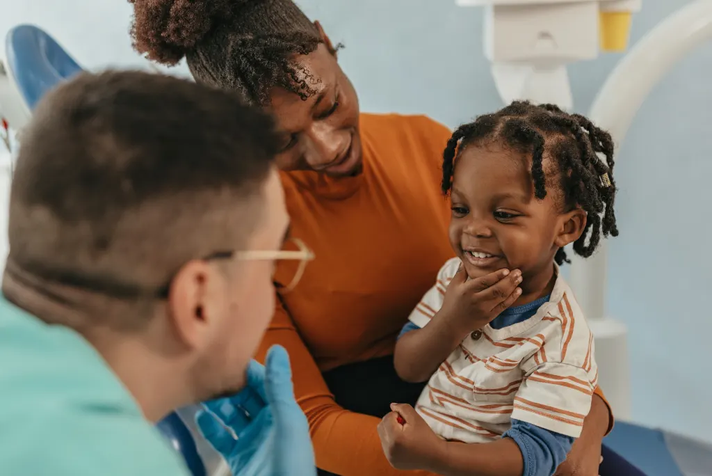 A young child sits on their mother's lap, smiling while interacting with a dentist in a clinic.