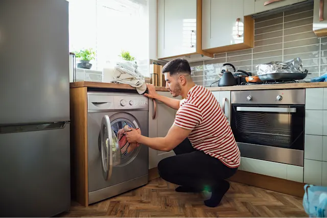 A man is loading laundry into a washing machine in a modern kitchen.
