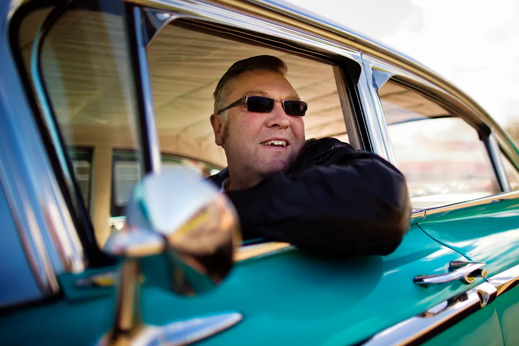 A man wearing sunglasses smiles while leaning out of the window of a classic teal car.