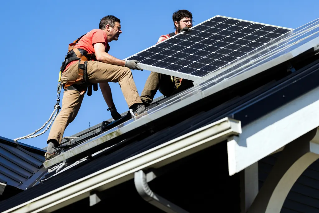 Two workers in safety gear are installing solar panels on a residential rooftop under a clear blue sky.