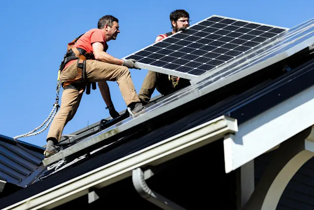 Two workers in safety gear are installing solar panels on a residential rooftop under a clear blue sky.
