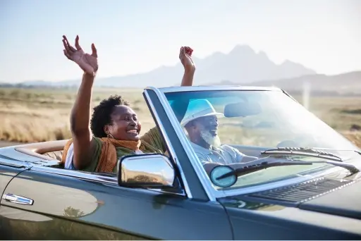 Man and woman driving in a older two seat convertible while the woman holds her hands up enjoying the ride