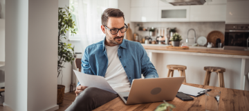 A man working remotely at home, focused on his laptop and reviewing documents at a kitchen table.