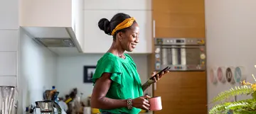 A smiling woman in a kitchen holding a mug and phone.
