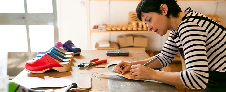 A craftsperson working on leather products in a workshop with colorful clogs and tools on display.
