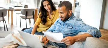 The image shows a couple sitting on a couch, reviewing documents together while using a laptop.