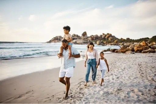 A family of four walking on the beach while the man carries a child on his shoulders and the woman holds hands with a child