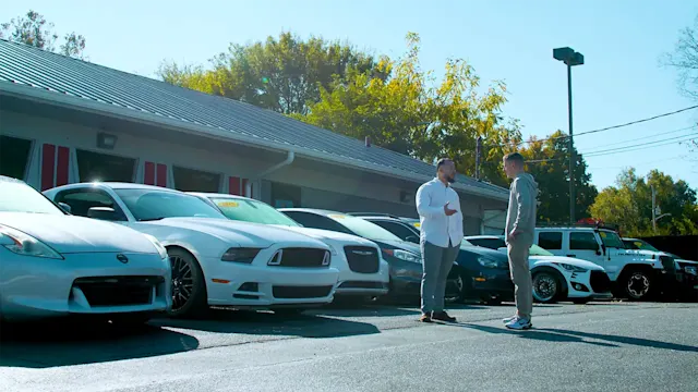 Two men having a conversation on a car dealers lot with parked cars in the background 