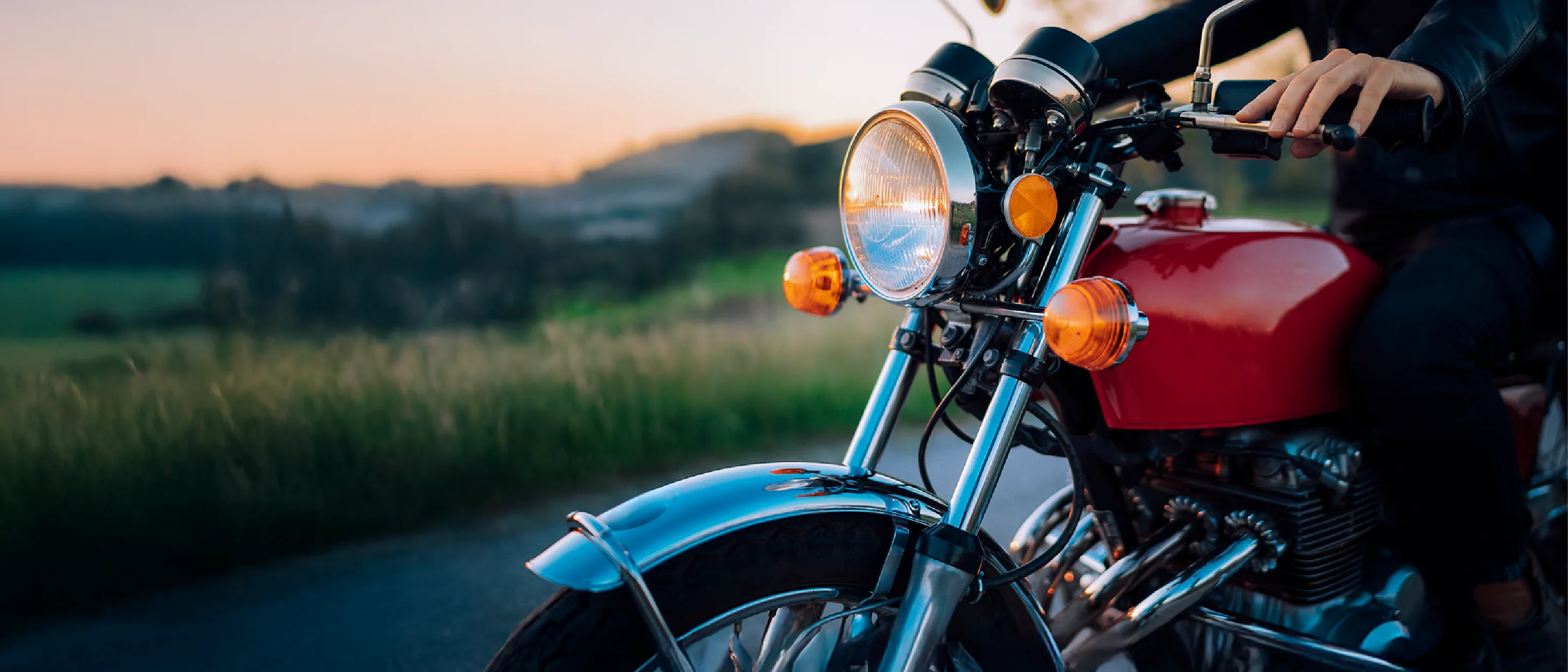 A person rides a classic red motorcycle down a scenic country road at sunset.