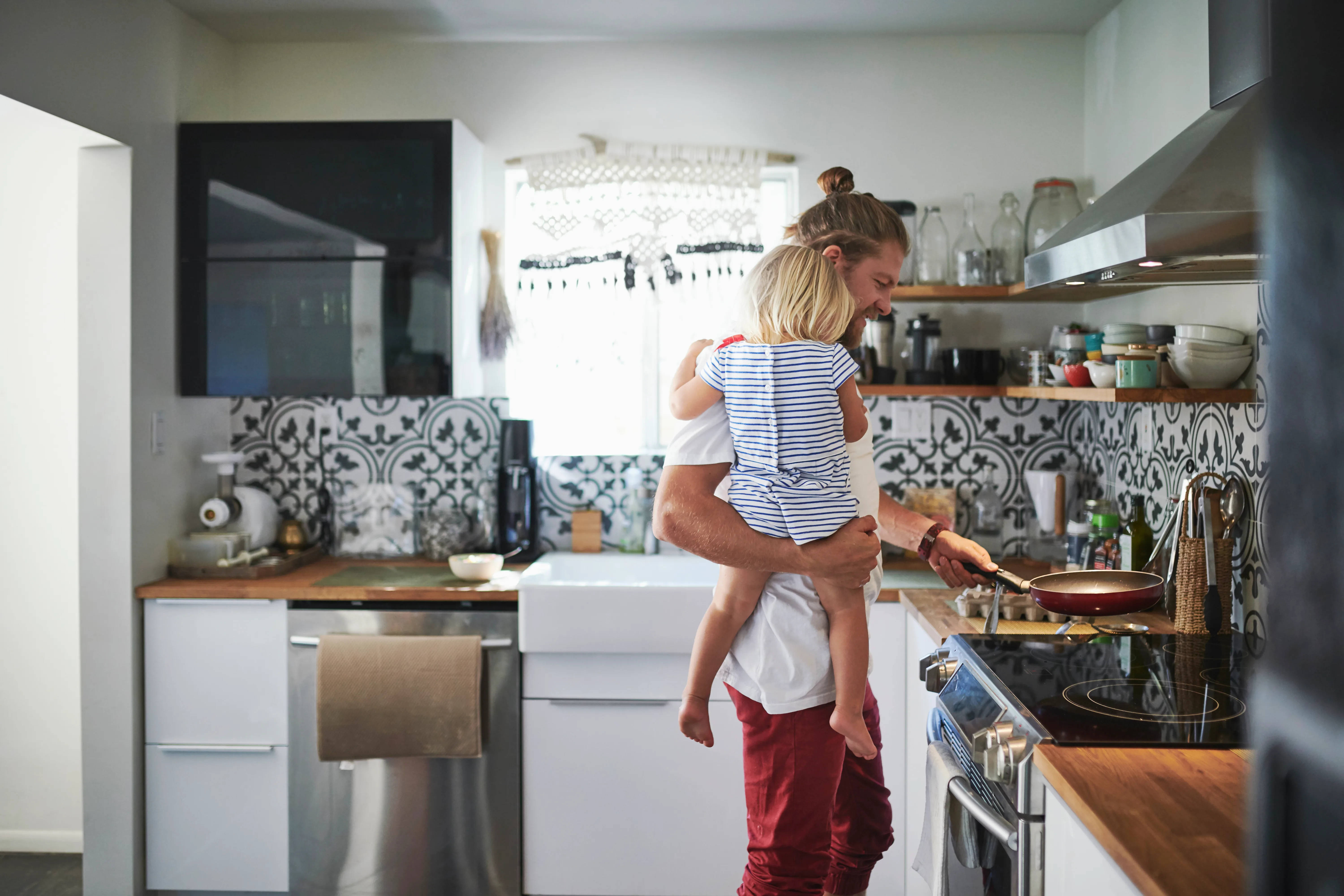 A dad is holding his child while cooking in the kitchen.