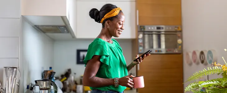 A smiling woman in a kitchen holding a mug and phone.