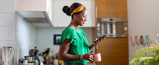 A smiling woman in a kitchen holding a mug and phone.