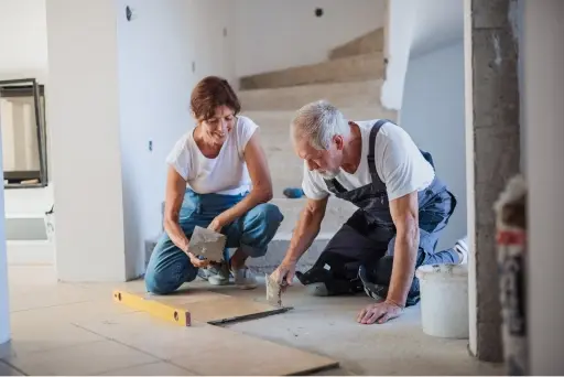 A man in overalls and a woman in jeans kneel on the floor to fix a section of the floor together