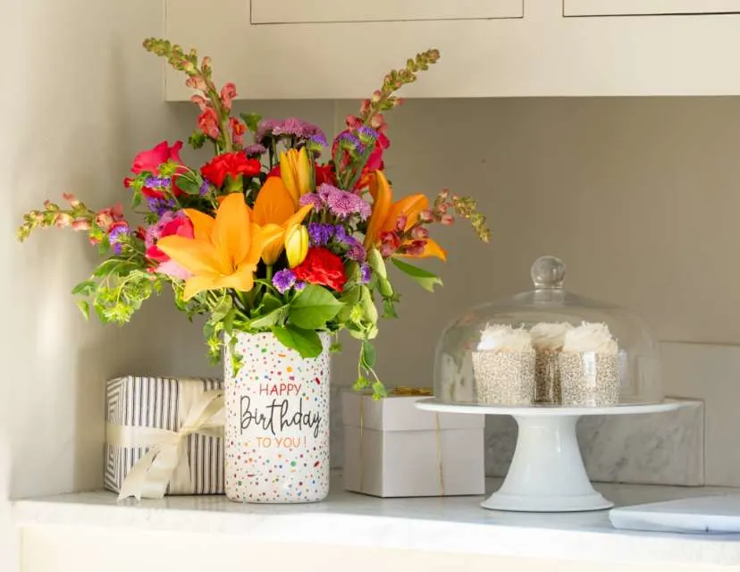 Off white kitchen counter featuring a colorful floral bouquet in a birthday vase, a vanilla cake inside a glass cake display and two wrapped presents