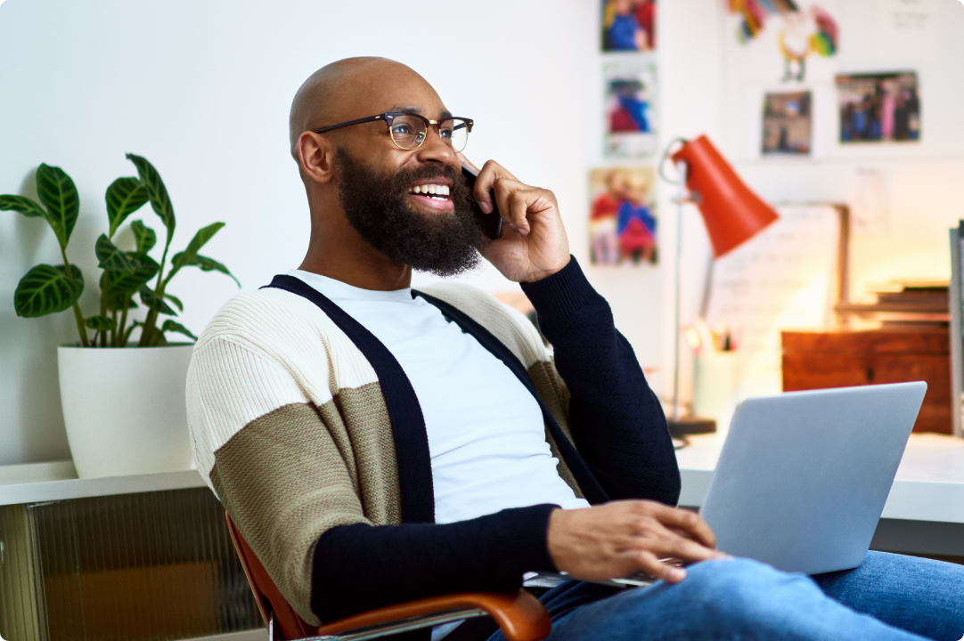 A Black person with a beard using a laptop and talking on the phone in a home office.