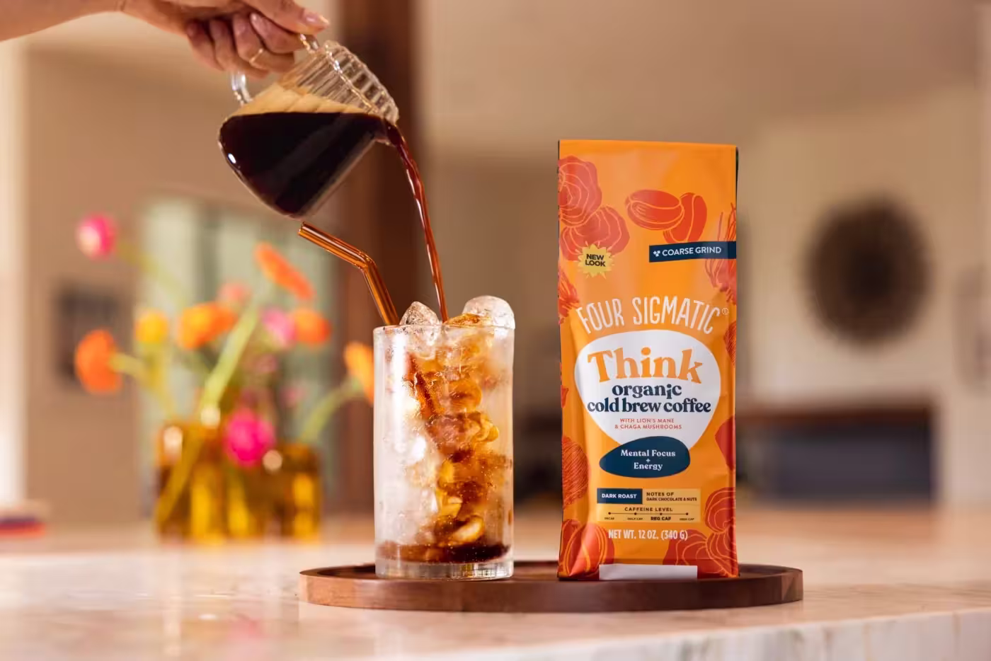 An orange 12-ounce bag of Four Sigmatic Think Organic Cold Brew Coffee with Lion's Mane and Chaga mushrooms. Next to it, dark coffee is poured from a glass pitcher into a tall glass filled with ice and a straw on a wooden coaster.
