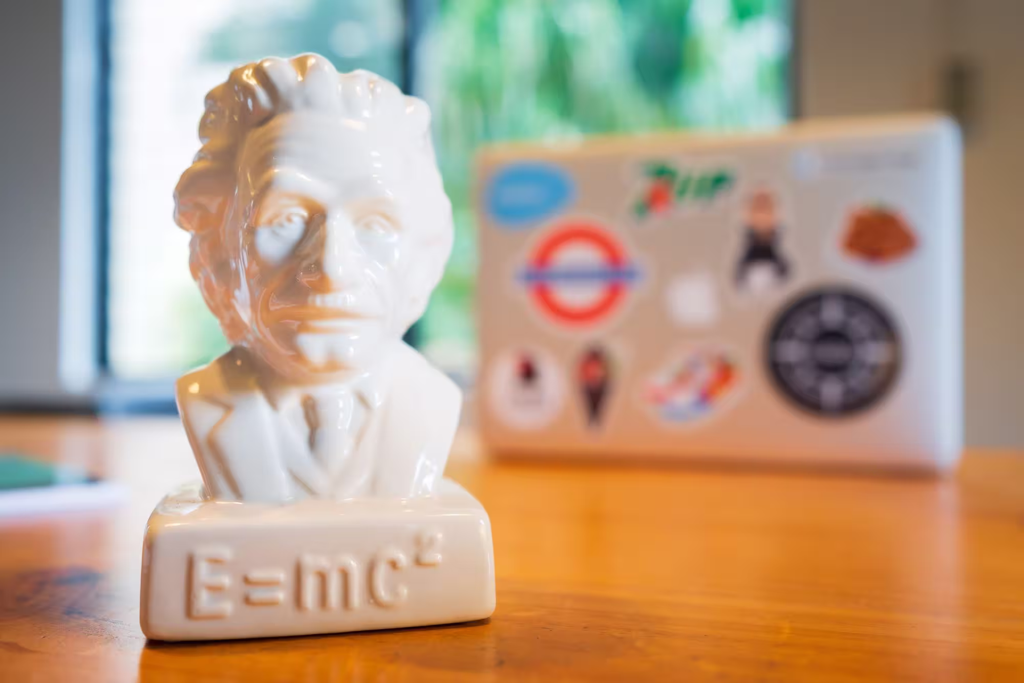 A small white ceramic bust of Albert Einstein featuring the equation $E=mc^2$ embossed on the base, sitting on a wooden desk with a blurred laptop in the background.