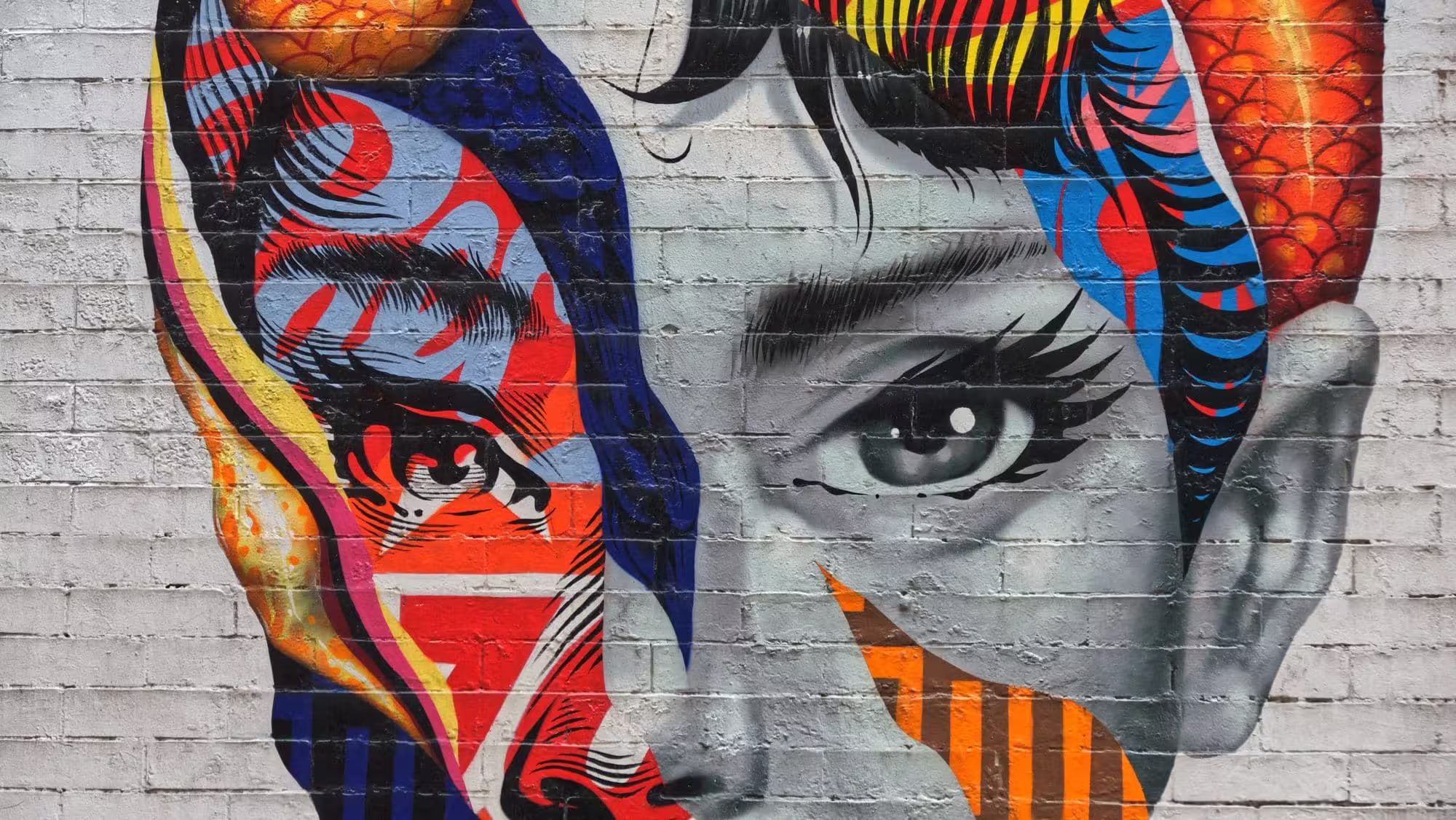 A street art mural of Audrey Hepburn painted on a white brick wall. Her face is split between a realistic grayscale portrait on the right and vibrant, colorful pop art patterns on the left.