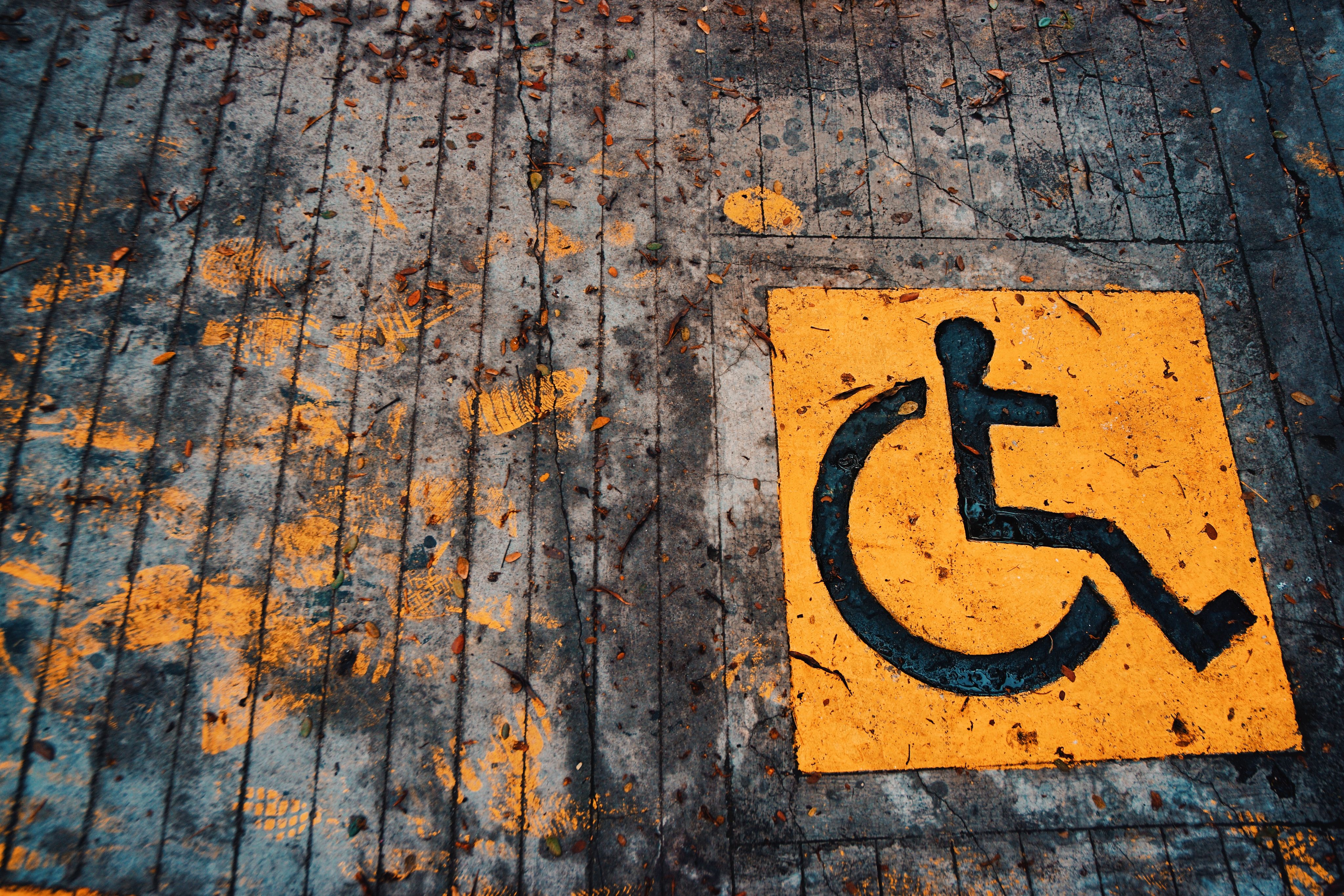 An international symbol of access wheelchair icon painted in black on a bright yellow square on concrete ground.
