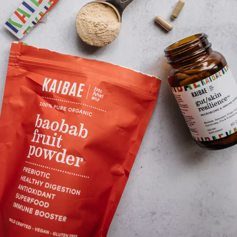 A flat lay of Kaibae wellness products on a light gray surface, featuring a large orange pouch of 100% Pure Organic Baobab Fruit Powder and an amber glass bottle of Gut/Skin Resilience dietary supplement capsules. A small metal scoop filled with the tan fruit powder and two loose capsules sit at the top.