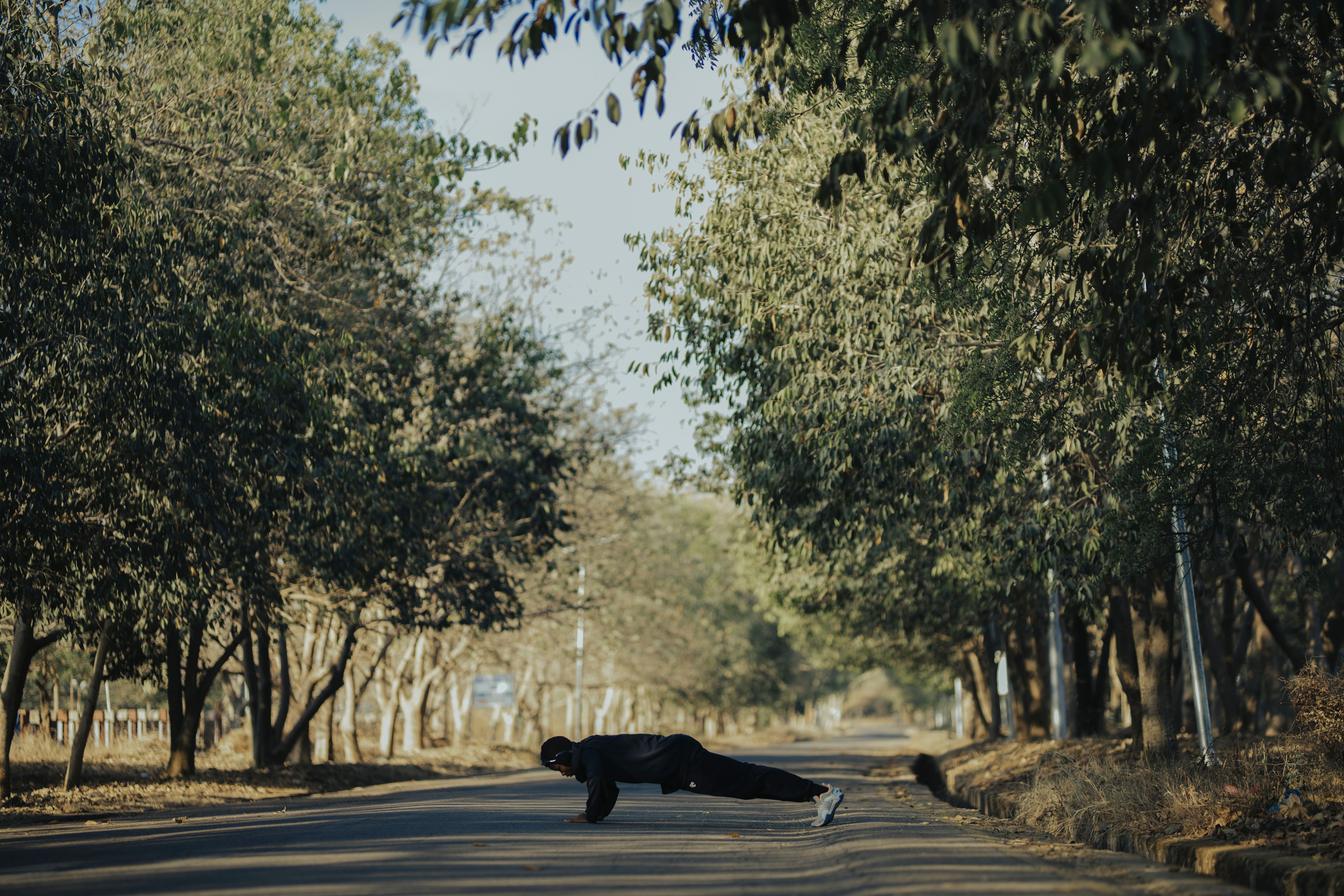 A person dressed in black athletic clothing performing a plank or push-up in the middle of an empty asphalt road. The road is heavily lined with tall green trees on both sides, casting soft shadows across the ground.