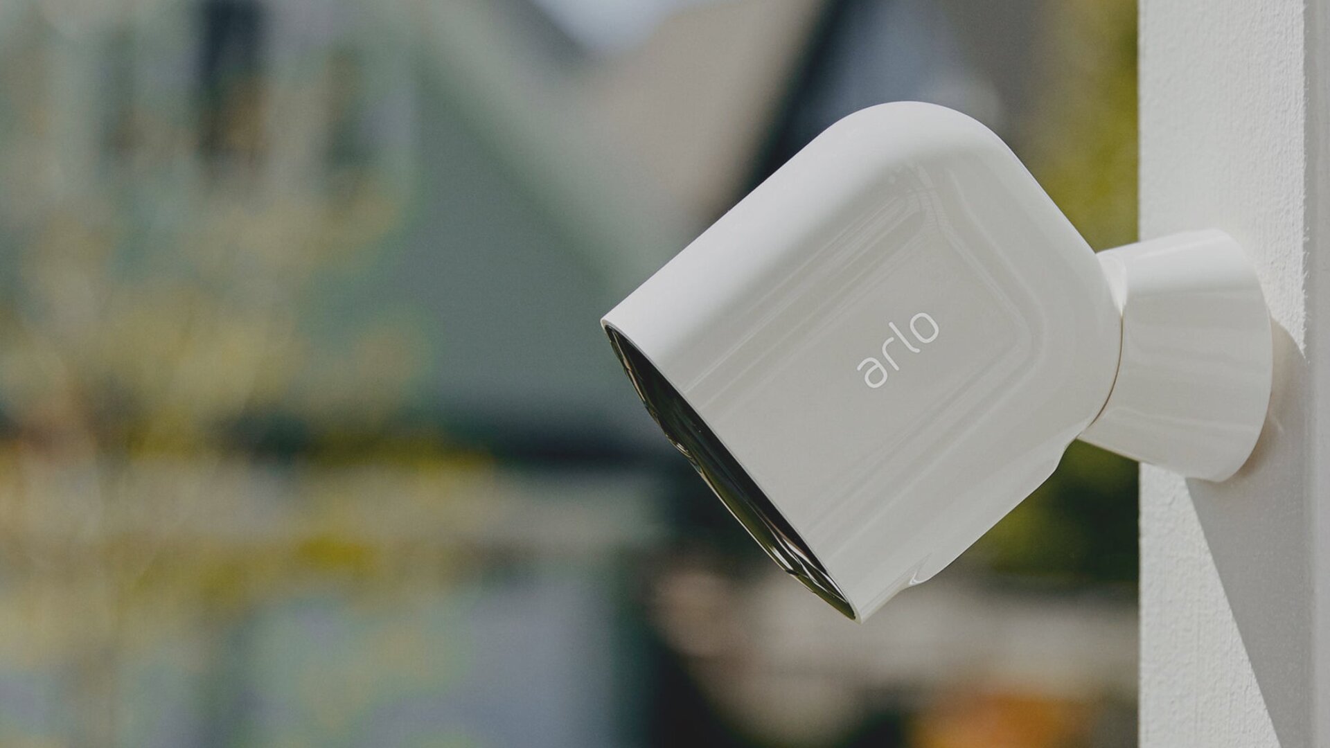A close-up of a glossy white Arlo smart security camera mounted onto a white exterior wall, angled downward with a softly blurred outdoor backyard scene in the background.