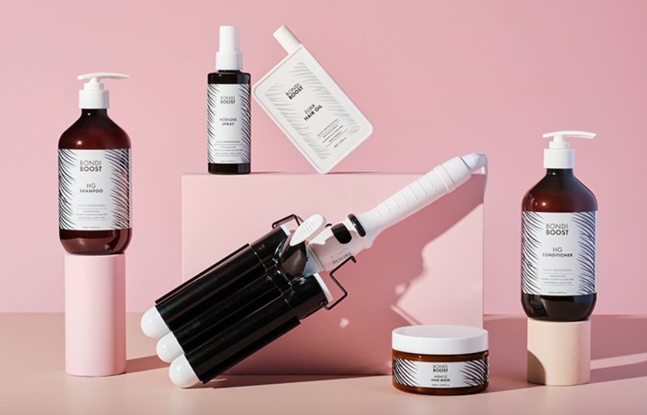 A collection of BondiBoost hair products displayed against a soft pink background. In the center sits a white and black triple-barrel hair waver styling tool, flanked by bottles of HG Shampoo, HG Conditioner, Intensive Spray, Elixir Hair Oil, and a jar of Miracle Hair Mask.