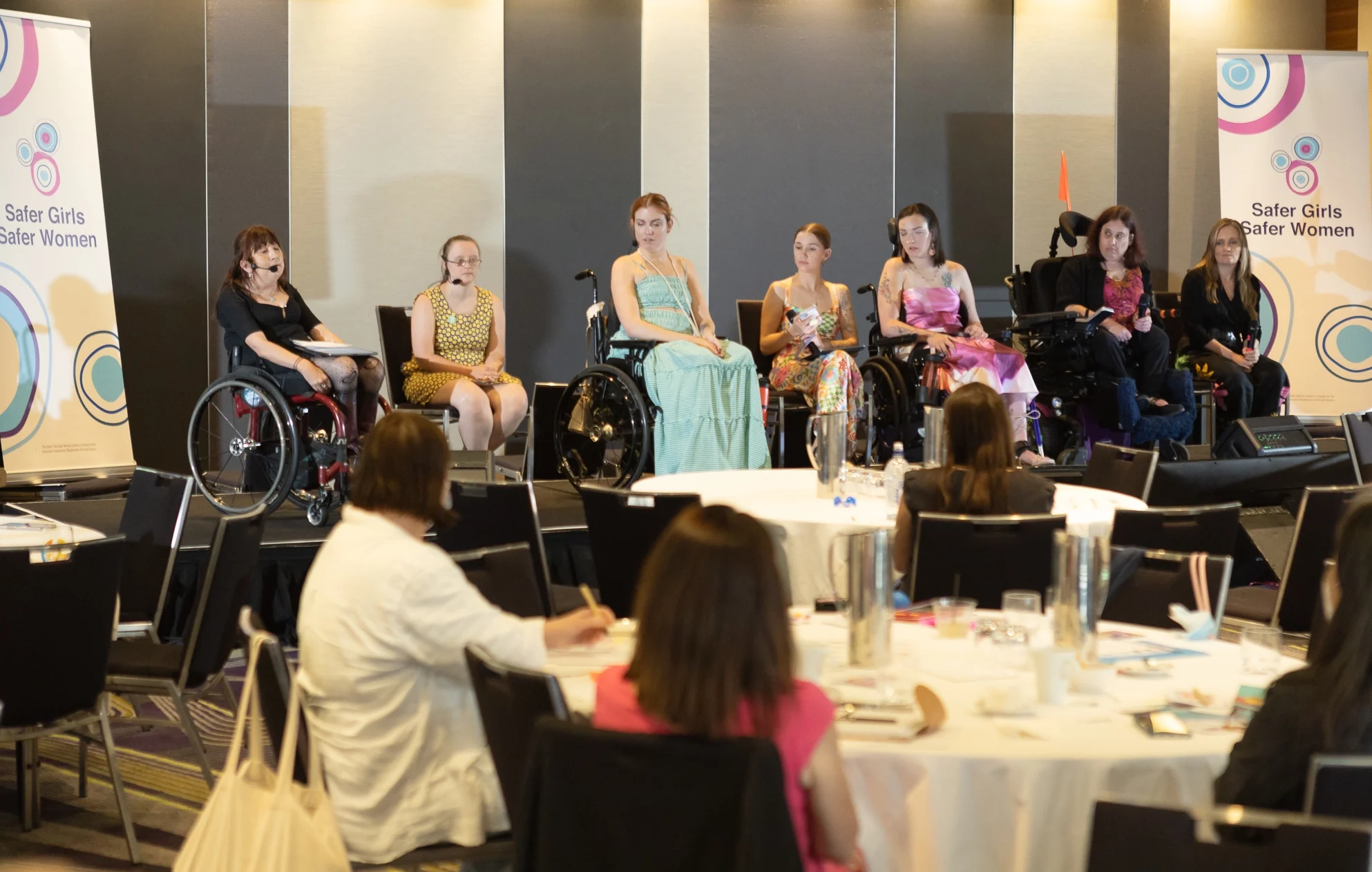 A panel of women, several in wheelchairs, sit at the front of a conference room beside a "Safer Girls Safer Women" banner, before an audience