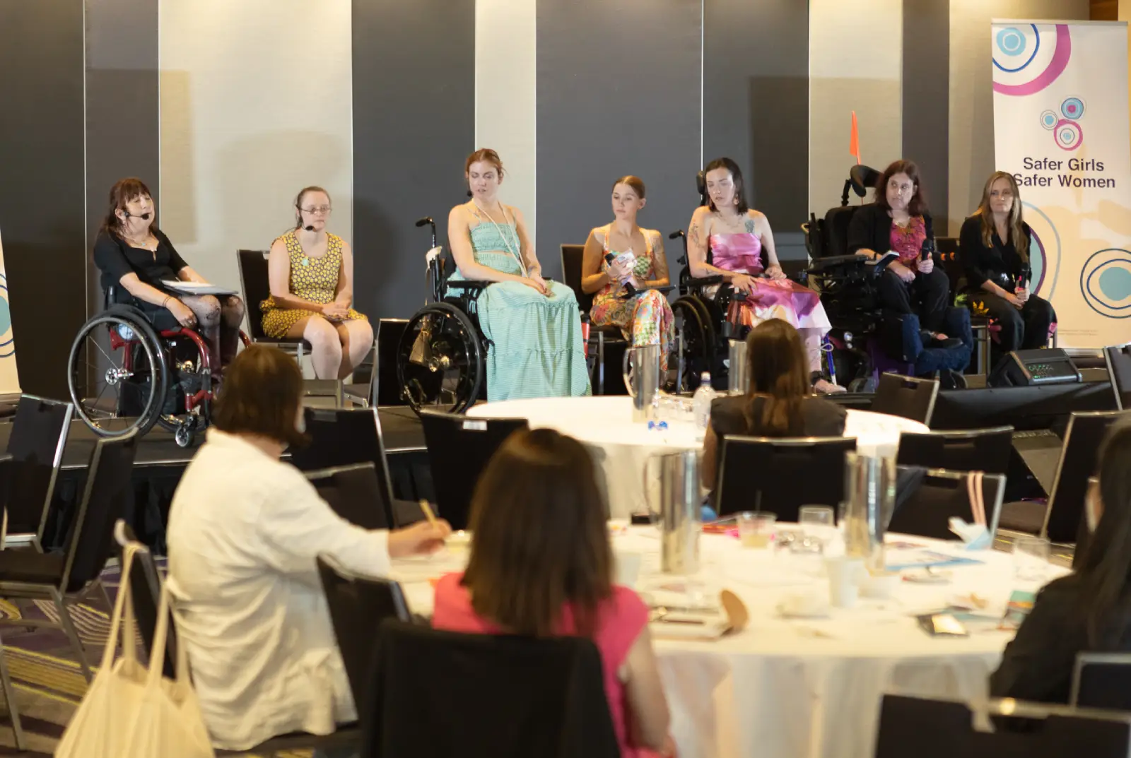 A panel of women, several in wheelchairs, sit at the front of a conference room beside a "Safer Girls Safer Women" banner, before an audience