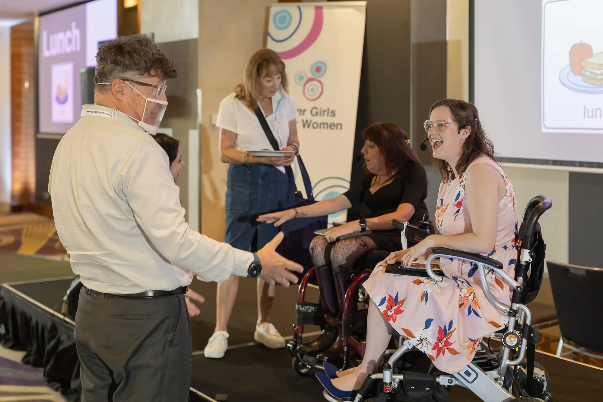 Four people interact near a "Safer Girls Safer Women" banner. An older man extends his hand to a laughing woman in a wheelchair, while another woman in a wheelchair listens intently. A woman stands in the background holding a tablet.