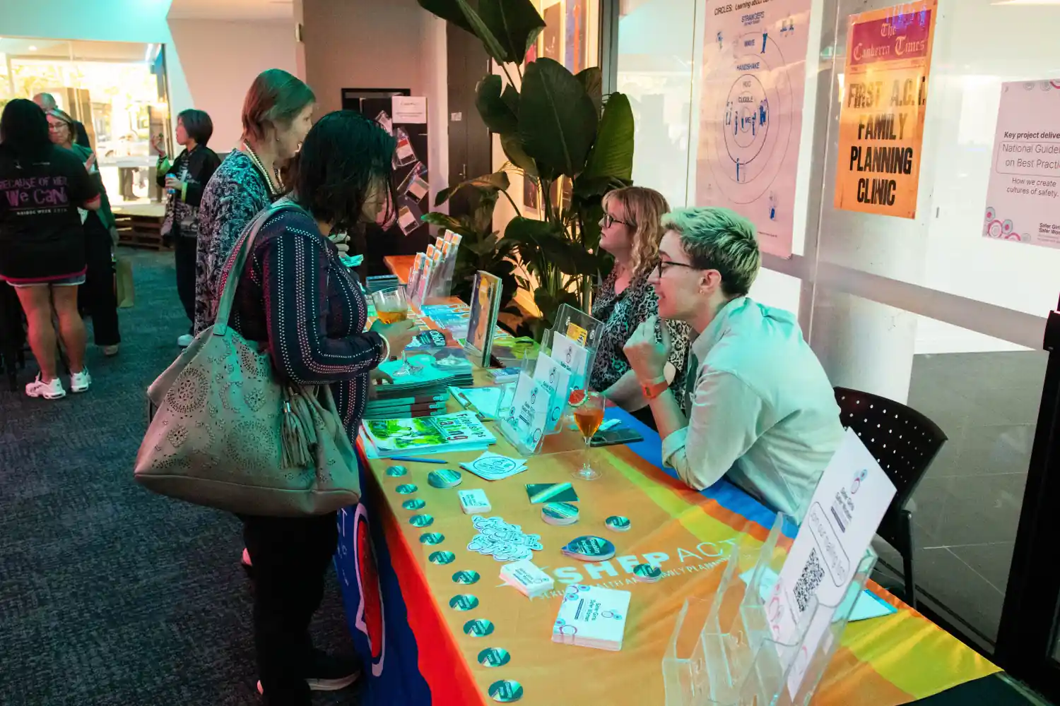 Visitors engage with representatives at the SHFPACT booth displaying educational materials and promotional items. Two attendees interact with two booth representatives, surrounded by other event paraphernalia and green plants.