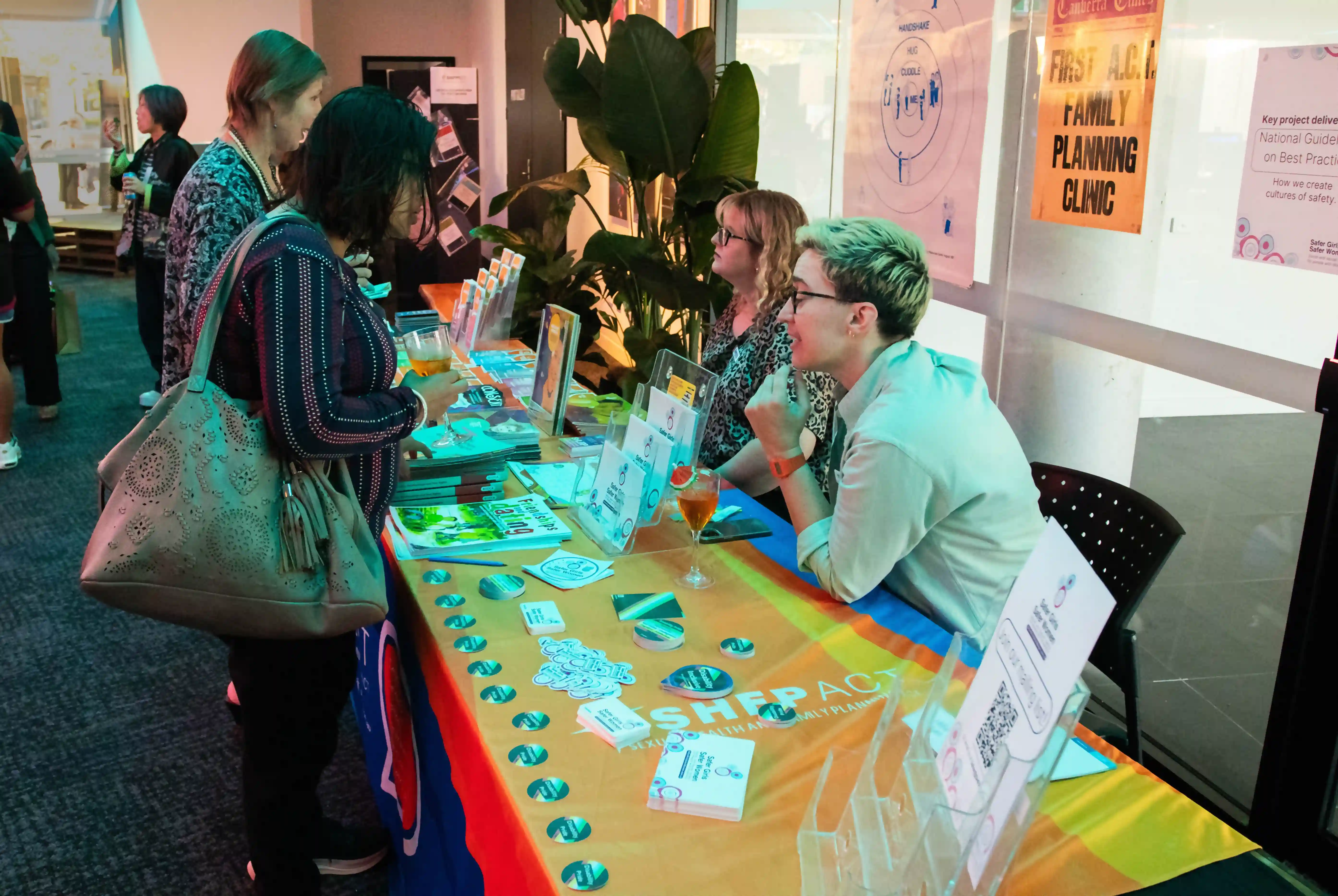 Visitors engage with representatives at the SHFPACT booth displaying educational materials and promotional items. Two attendees interact with two booth representatives, surrounded by other event paraphernalia and green plants.