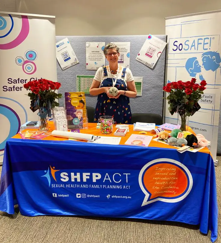 A woman stands behind an SHFPACT booth at a conference, smiling and holding a plush koala. The table is covered with a blue cloth featuring the organization’s logo and displays various sexual health and rights resources. Background posters promote the SoSAFE! program.