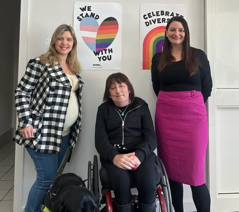 Three women are posing together in front of posters that read "We Stand With You" and "Celebrate Diversity". One woman, standing on the left, is wearing a black-and-white checkered coat. The woman in the middle is seated in a wheelchair with a service dog by her side. The woman on the right is wearing a black top and a bright pink skirt.