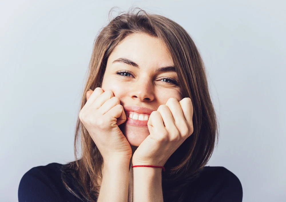 Eine Frau, die in in die Kamera lächelt und ihr Gesicht auf ihren Händen abstützt.