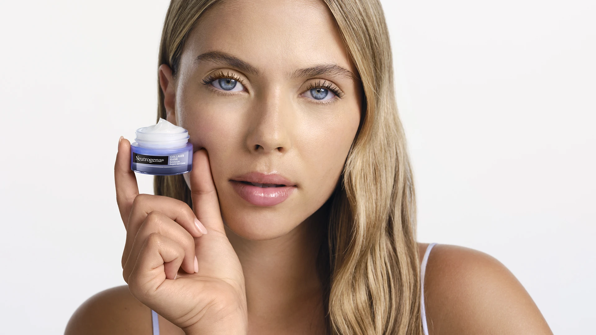 Blonde Frau hält die Neutrogena® Collagen Bank Belebende Augen-Gel-Creme in der Hand neben ihrem Gesicht.