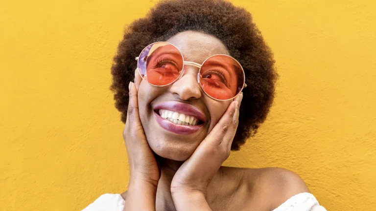 A Black woman in large, tinted glasses smiles and holds her cheeks in front of a yellow wall 