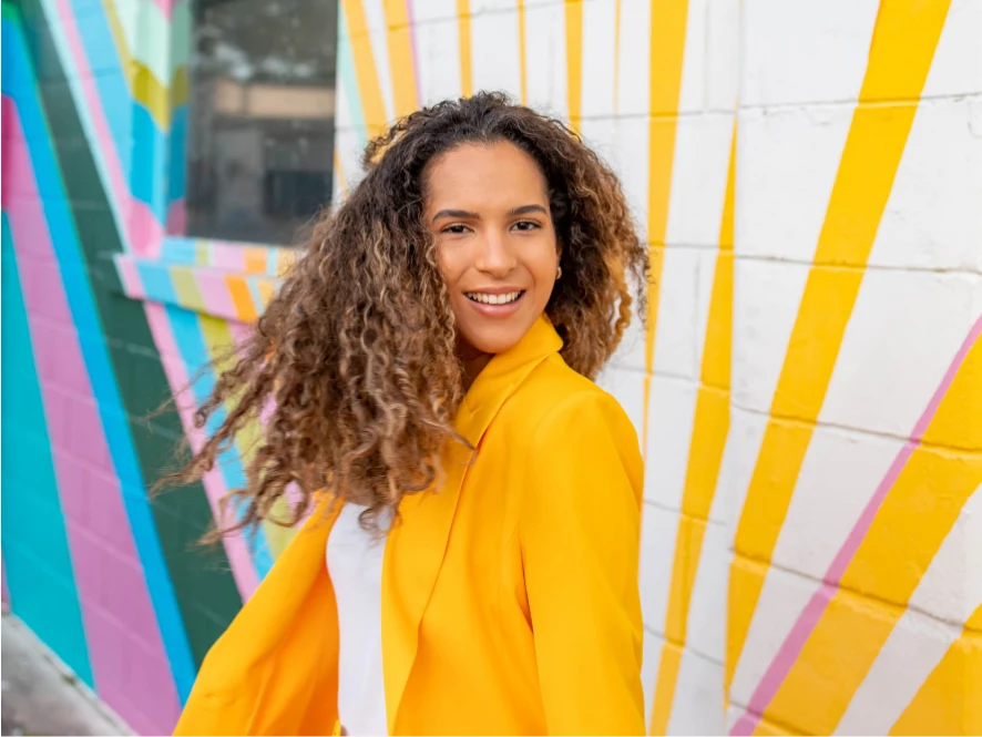 Jomely Breton sonriendo y volteando a la cámara, usando un saco amarillo brillante, una pared colorida de fondo