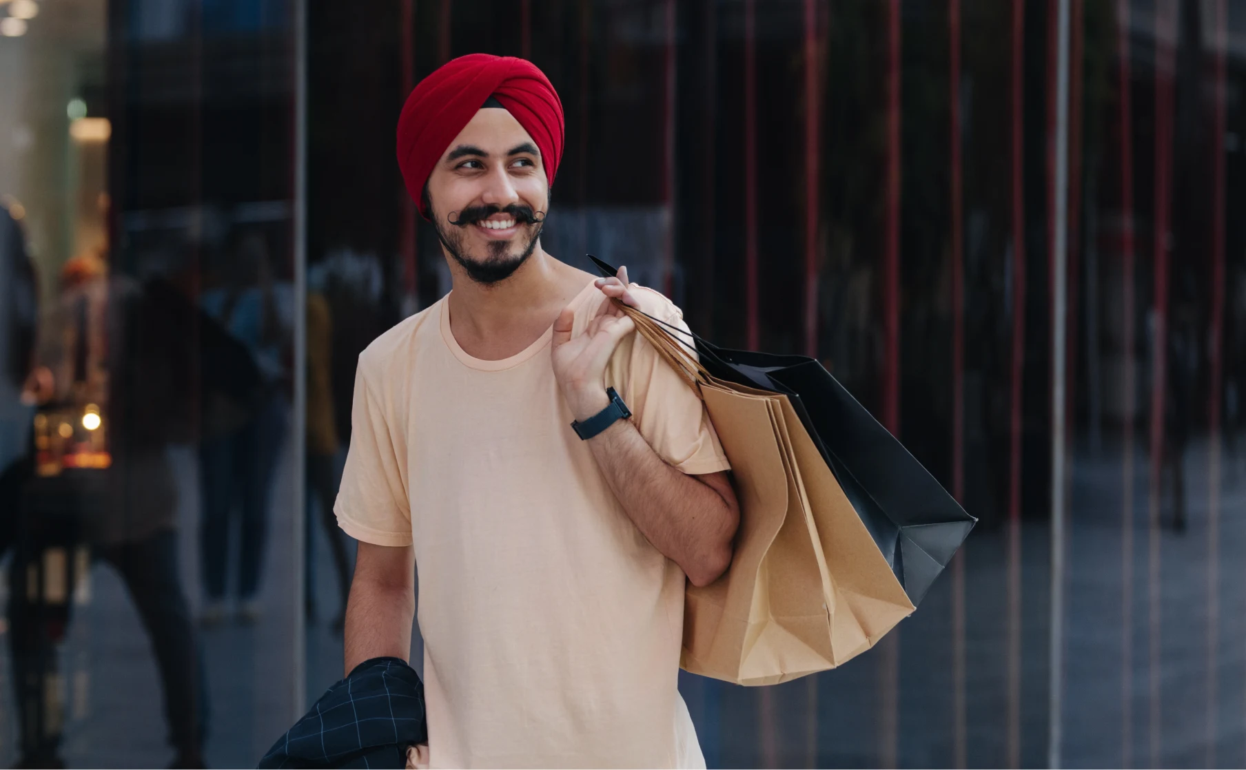 South Asian man in a red head wrap smiles. He holds a brown shopping back and cell phone. Dark blue blazer draped over his arm. Muted storefront in the background. Tree shadows fall on the facade. Green plants in front.