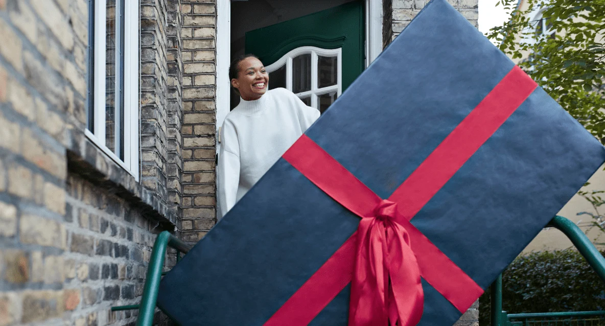 A smiling Black woman accepts a large gift wrapped in blue paper with a big red bow, at the front door of her brick home