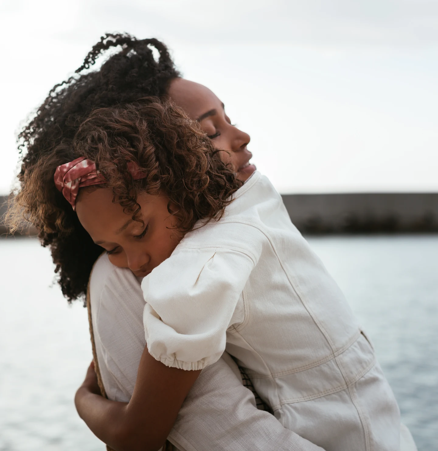 Mother and daughter wearing white dresses hugging in front of a body of water.