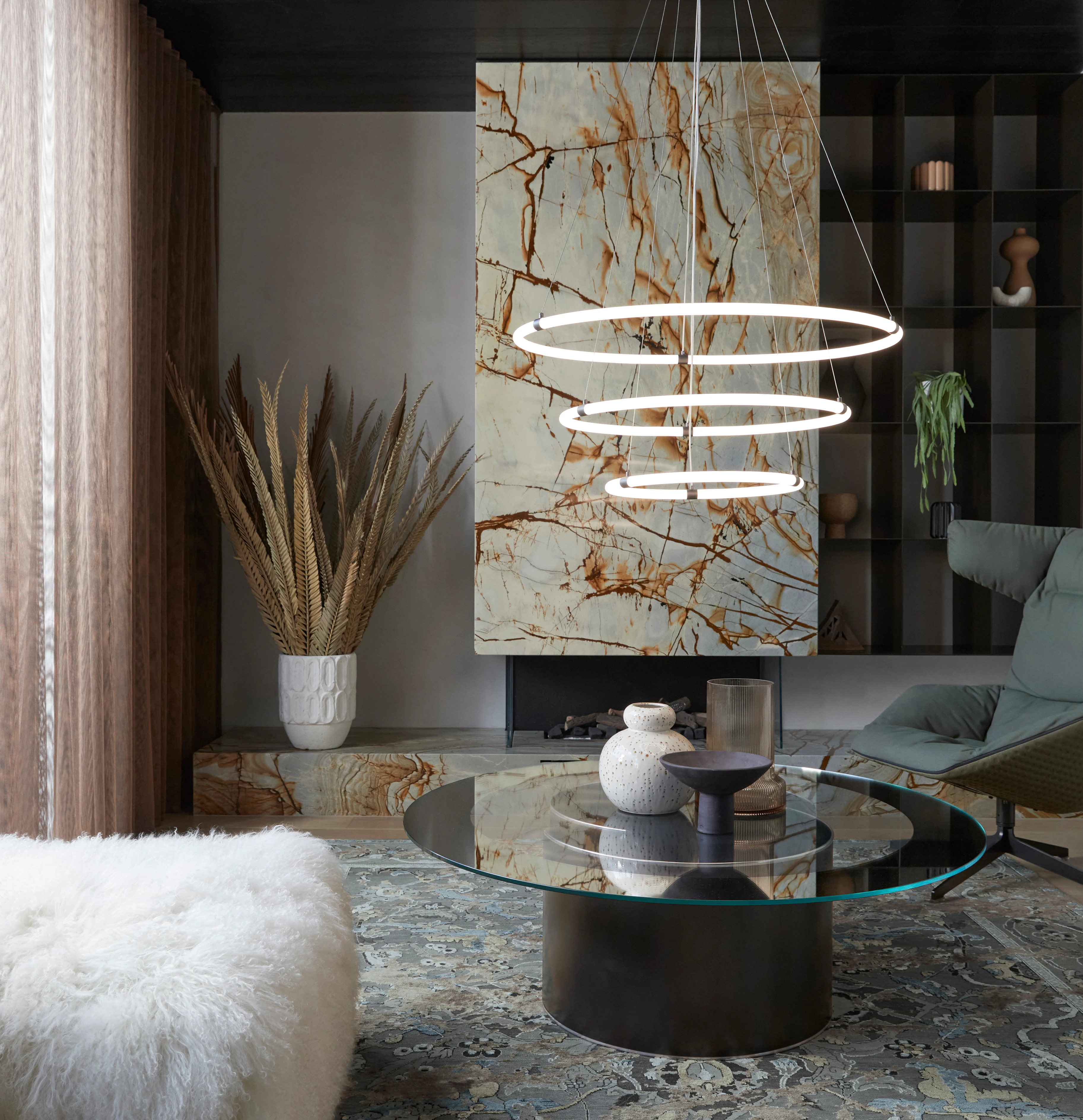 Image of a modern living room. To the left, tan curtains and a fluffy, white seat. In the left-hand corner, dried feathers in a large white vase. In the middle, a circular mirror tabled with three vases on top. A marble painting sits in the background and, above the table, a cylindrical light fixture made up of three circles. To the right, a bookcase and a forest green reclining chair.