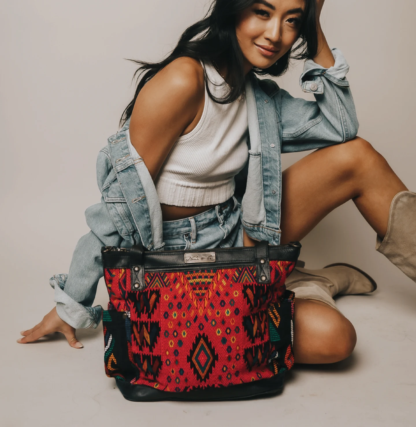 East Asian female model in white knitted tank top, jean jacket and jean skirt sits and smiles behind a red Nena & Co. handbag