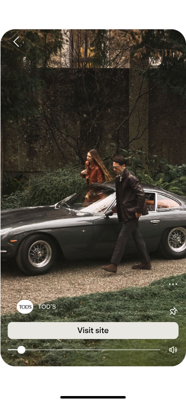 A man and woman in stylish attire walk past a vintage car on a gravel path, surrounded by lush greenery, conveying a sense of elegance and nostalgia.